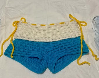 Puede incluir: Shorts tejidos a mano en crochet con un diseño de dos tonos. La banda superior es de color crema, con costuras y cordones amarillos. Los shorts son de un azul vibrante. La prenda está hecha de un hilo suave y texturizado.