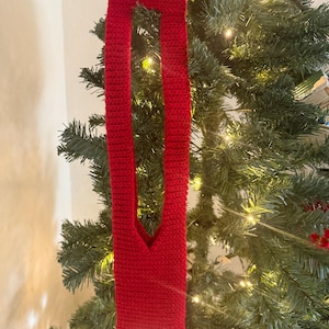 May include: A red, crocheted wine bottle holder with a long, rectangular shape and a central opening. The holder is hanging on a Christmas tree with green needles and small, twinkling lights.
