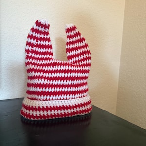 May include: A handmade crochet hat in red and white stripes. The hat has a wide brim and two upright, ear-like extensions. The hat is sitting on a dark surface, with a neutral-colored wall in the background.