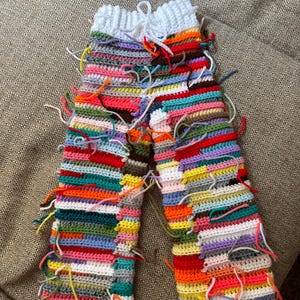 Puede incluir: Pantalones de ganchillo hechos a mano con un vibrante diseño de patchwork. Los pantalones muestran rayas horizontales en una variedad de colores, incluyendo rojo, azul, amarillo y verde. El ganchillo blanco forma la cintura. Los extremos sueltos del hilo añaden textura.