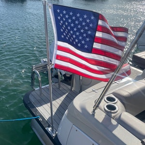Könnte beinhalten: Ein weißes Boot mit einem Holzdeck und einem Edelstahlflaggenmast, an dem eine amerikanische Flagge befestigt ist. Das Boot liegt in einem Yachthafen mit anderen Booten im Hintergrund.