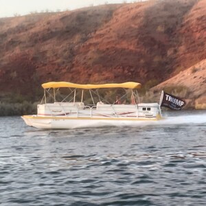 Puede incluir: Un barco pontón blanco con una cubierta amarilla se mueve a través de una masa de agua. Una bandera negra con la palabra "TRUMP" ondea en la parte trasera del barco.