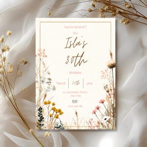 May include: A cream-colored invitation card with gold text and a gold border. The text reads "You're Invited!! It's Isla's 30th Birthday March 25th 3PM". Floral illustrations in yellow, pink, and white decorate the edges.