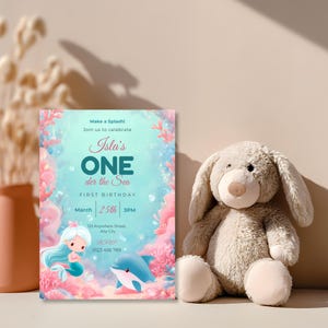 Baby girl’s mermaid first birthday invitation standing beside a soft plush bunny, with aqua water, pink coral and a cute dolphin.