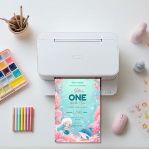 Printable Oneder the Sea first birthday invitation coming out of a home printer beside pastel markers and a watercolor paint palette.
