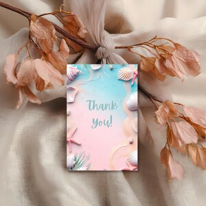 May include: A pastel-colored greeting card with a beach theme. The card features seashells, starfish, and the words "Thank you!" in a soft, elegant font. The background has a gradient of blue and pink, evoking a seaside scene.