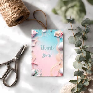 May include: A rectangular gift tag with a beach-themed design. The tag features seashells, starfish, and the words "Thank you!" against a pastel pink and blue background. A roll of twine and scissors are visible.