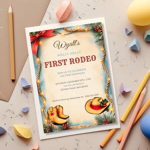 Printed Holly Jolly First Rodeo invitation on a desk with colored pencils, showing the dusty blue, tan and barn red cowboy Christmas design