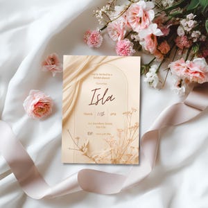 May include: A bridal shower invitation with the name "Isla" in elegant script. The invitation features a beige background with floral and ribbon accents. The text includes the date, time, and RSVP information. Soft pink flowers and a satin ribbon surround the invitation.