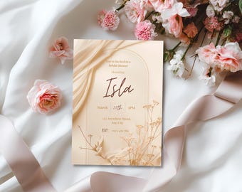 May include: A bridal shower invitation with the name "Isla" in elegant script. The invitation features a beige background with floral and ribbon accents. The text includes the date, time, and RSVP information. Soft pink flowers and a satin ribbon surround the invitation.