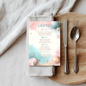 May include: A menu card with a beach theme, featuring the text "Let's Eat" in elegant script. The menu lists starters, mains, drinks, and sweet treats, with seashell and starfish illustrations. A knife, fork, and spoon are on a wooden board.