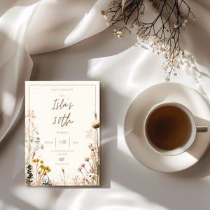 May include: An invitation card with floral illustrations and the text "Isla's 30th Birthday." A cup of tea on a saucer is placed next to the card. The background is a white, silky fabric.