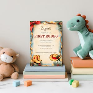 Western Christmas 1st birthday invitation for a baby boy displayed on a nursery shelf with stacked books, a plush dinosaur and a teddy bear