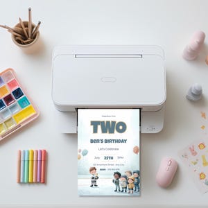 Printed ice hockey birthday invitation coming out of a printer, featuring watercolor children celebrating on ice rink with “Slapshot Into TWO” title