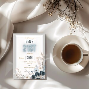 May include: A birthday invitation with the text "BEN'S 21ST" in light blue. The invitation includes the date, time, and location. Floral accents in white and blue decorate the border. A cup of tea sits on a saucer next to the invitation.