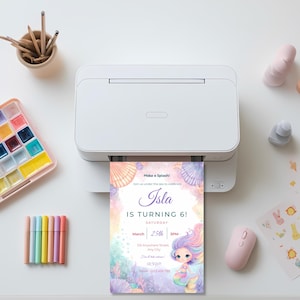 Printable mermaid birthday invitation coming out of a home printer, surrounded by pastel markers and a watercolor paint palette.