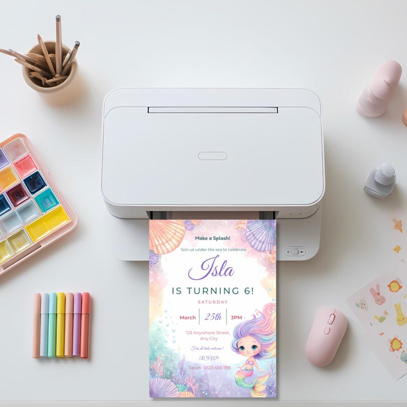 Printable mermaid birthday invitation coming out of a home printer, surrounded by pastel markers and a watercolor paint palette.
