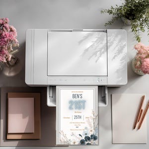 May include: A white printer printing a birthday invitation. The invitation reads "Ben's 21st Birthday" with the date and time. The invitation has a floral design with blue and white flowers. Pink flowers and pens are on the table.