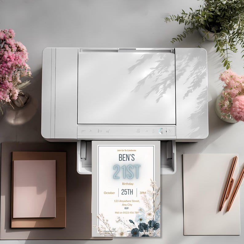 May include: A white printer printing a birthday invitation. The invitation reads "Ben's 21st Birthday" with the date and time. The invitation has a floral design with blue and white flowers. Pink flowers and pens are on the table.