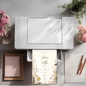 May include: A white printer printing a birthday invitation. The invitation has a floral design with the text "Isla's 30th Birthday". Other items on the table include stationery, flowers, and pens.