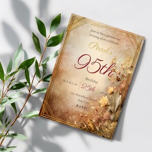 95th birthday invitation template in Art Deco amber and gold displayed as a printed card tilted at an angle, with large green tropical leaves casting soft shadows behind it