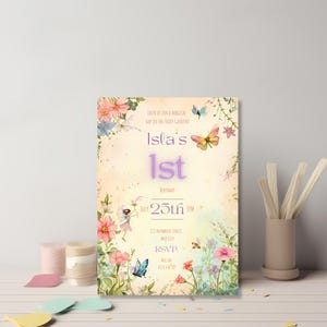 Fairy garden birthday invitation template standing on a white shelf surrounded by pink candles and craft supplies, featuring watercolor wildflowers, butterflies and a whimsical fairy illustration on a warm peach background