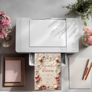 May include: A white printer printing a romantic dinner invitation. The invitation features a cream background with red hearts, candles, and the text "Romantic Dinner". Pink flowers and pens are also visible.