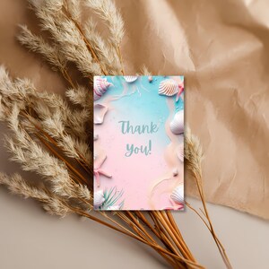 May include: A greeting card with a beach theme. The card features a pastel pink and blue background with seashells, starfish, and the text "Thank you!" in a script font. Dried grasses and a tan paper background surround the card.