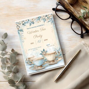 Editable winter tea party invitation on a wood desk with glasses and a pen, elegant blue and gold snowflake design ready to personalize in Canva.