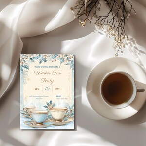 Editable winter tea party invitation displayed beside a cup of tea, elegant blue and gold design with gold teacups and snowflake accents.