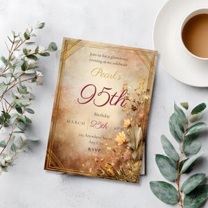 95th birthday invitation template in Art Deco amber and gold with dried floral botanicals, displayed as a printed card on a white surface beside eucalyptus stems and a cup of tea