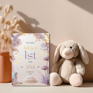 Fairy first birthday invitation printable template displayed beside soft toy bunny in warm styled scene showing enchanted purple fairy mushroom lavender design for baby girls first birthday
