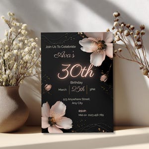 Elegant black birthday invitation template with blush florals and gold accents, shown leaning against a vase with dried flowers, editable in Canva