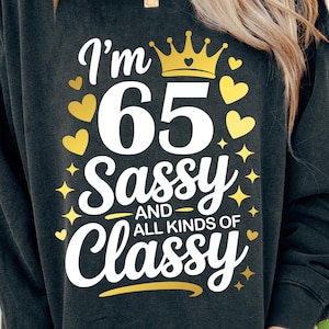 May include: Dark grey sweatshirt with the text "I'm 65 Sassy and All Kinds of Classy" in white script. The number 65 is large and white, with a gold crown above it. Gold hearts and stars are scattered around the text.