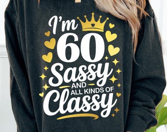 以下が含まれることがあります： 「I'm 60 Sassy and All Kinds of Classy」の文字が白と金でプリントされた濃いグレーのスウェットシャツ。デザインには金の王冠、ハート、星が含まれています。記念日の誕生日のためのカジュアルで快適なトップス。