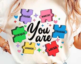 May include: A white t-shirt with a colorful puzzle design that says "You are" in the center. The puzzle pieces have the words "valued", "worthy", "unique", "treasured", "chosen", and "loved" on them.