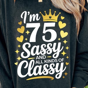 May include: Dark gray sweatshirt with the text "I'm 75 Sassy and All Kinds of Classy" in white script. The design includes a gold crown, hearts, and stars. A casual, celebratory garment.