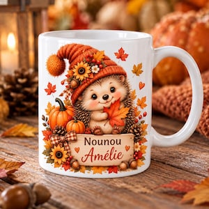 Peut inclure: Un mug en céramique blanche orné d'un hérisson portant un bonnet en tricot orange, entouré de feuilles d'automne, de citrouilles, de tournesols et de glands. Le mug affiche le texte Nounou Amélie sur un panneau en bois avec des petits cœurs.