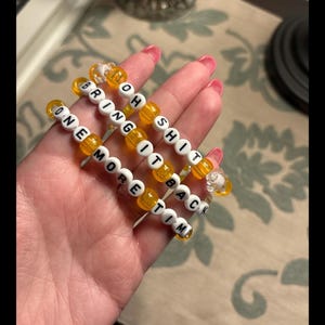 Peut inclure: Trois bracelets en perles jaunes avec des perles de lettres blanches qui épellent la phrase "Oh Shit Bring It Back One More Time".