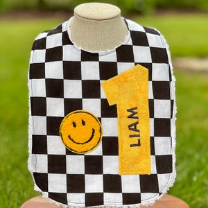 May include: A black and white checkered baby bib with a yellow smiley face and a yellow number one with the name Liam on it.