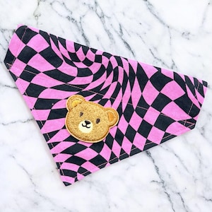 May include: A pink and black checkered pet bandana with a wavy pattern. The bandana features a brown teddy bear patch with a smiling face. The fabric is a soft material, perfect for pets.