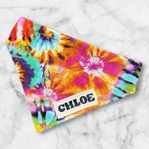 May include: A colorful tie-dye dog bandana with a vibrant pattern of orange, pink, green, and blue hues. The bandana features the name "CHLOE" on a small, rectangular label. The fabric is folded into a triangle shape.
