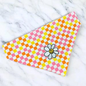 May include: Pink, orange, and yellow checkered bandana with a white daisy embroidered with a yellow smiley face.