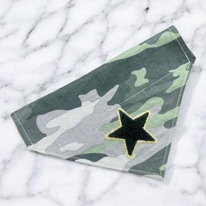 May include: A camouflage-patterned pet bandana in shades of green, gray, and white. A black star with gold glitter trim is sewn onto the bandana. The bandana is folded in a triangle shape.
