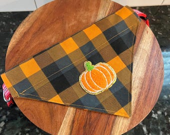 May include: A black and orange plaid dog bandana with a felt pumpkin applique. The bandana has a red and white adjustable strap.