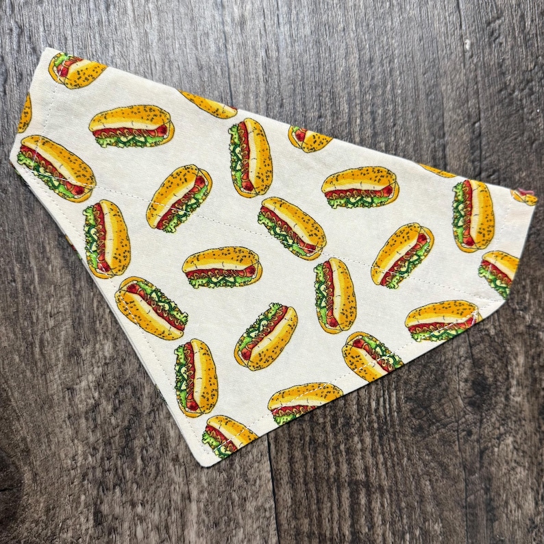 May include: A white cotton dog bandana with a repeating pattern of yellow hot dogs with mustard and relish on a white background.