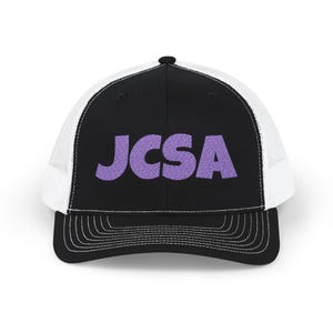 Puede incluir: Gorra de camionero en blanco y negro con las letras "JCSA" bordadas en morado. La gorra presenta un panel frontal negro, una parte trasera de malla blanca y una visera curva con costuras blancas.