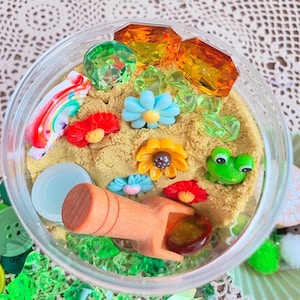 May include: A clear plastic container filled with sand, colourful gems, and small plastic flowers. A wooden rake and a green frog figurine are also inside. The gems are orange, green, and clear. The flowers are red, yellow, and blue.