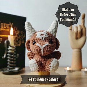 Può includere: Un giocattolo di peluche in crochet marrone e bianco a forma di toro con le corna. Il giocattolo è seduto su una base di legno con il testo "24 Couleurs / Colors" su di essa. Il testo "Make to Order / Sur Commande" è visibile sullo sfondo.