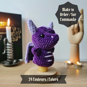 Peut inclure: Peluche de dragon en crochet violet avec une crinière et des ailes violettes claires. Le jouet est posé sur une base en bois avec le texte "24 Couleurs / Colors" et "Make to Order / Sur Commande" sur un fond blanc.
